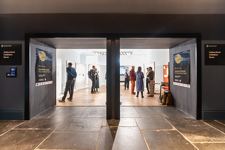 A group of people stands and talks inside the brightly lit Anthea Bond  Exhibition Room, viewed through two wide entryways from a darker corridor with slate flooring. Posters and signs are displayed on both sides of the entrances.