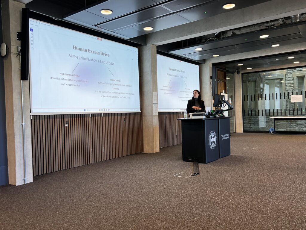 A woman stands at a lectern giving a presentation in a modern lecture theatre. The slide on the screen is titled "Human Excess Drive" and discusses animals and behavioural perception. The room is spacious with wood panelling and large windows.