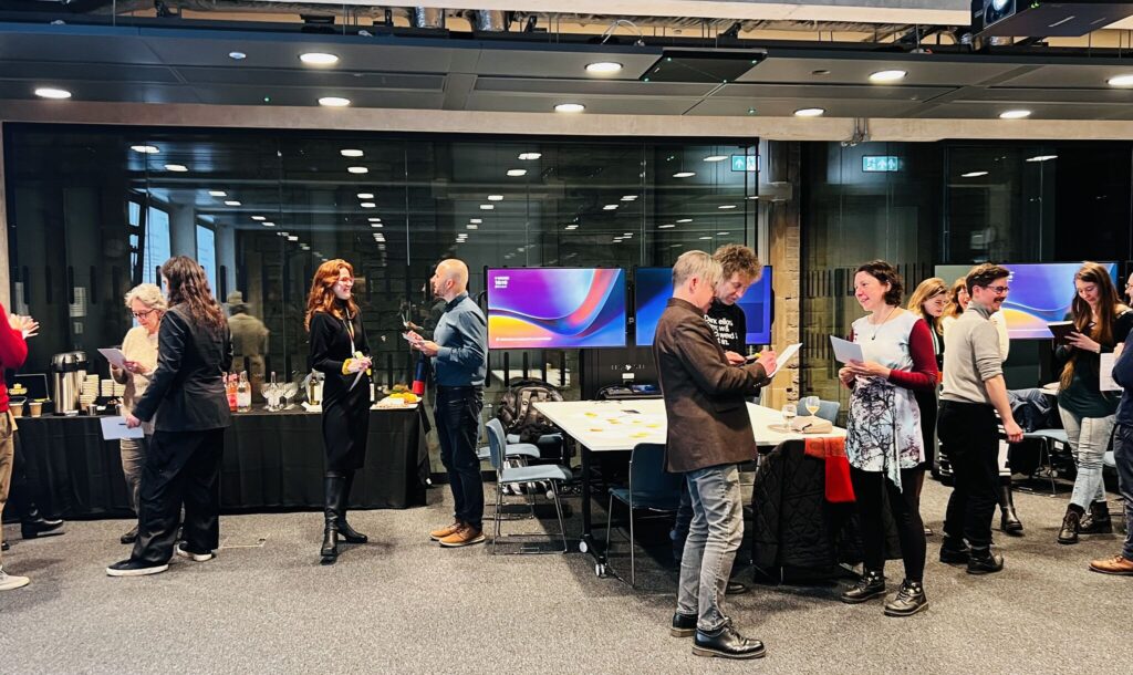 A group of people stand and chat in pairs or small groups in a modern conference room with large windows, digital screens, and tables with papers and drinks. The setting appears to be a networking or workshop event.