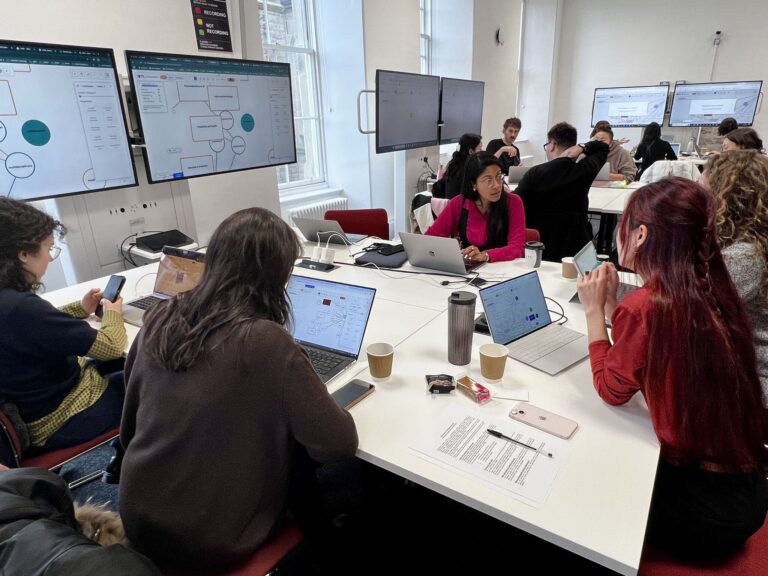 A group of people sit around tables with laptops, collaborating and discussing in a bright room. Large screens display diagrams and notes. Papers, cups, and notebooks are on the tables. The atmosphere is focused and engaged.
