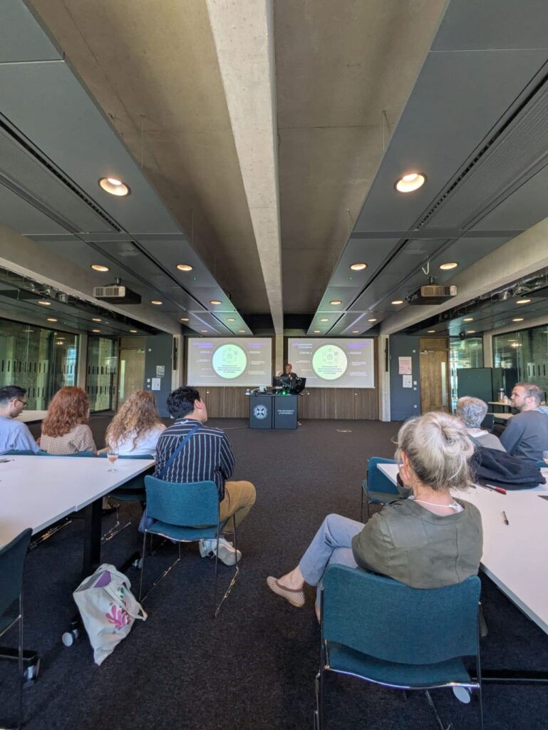 A group of people sit in a modern classroom, facing a presenter at a lectern. Two large screens behind the presenter display the same presentation slide with a circular diagram and purple sections.