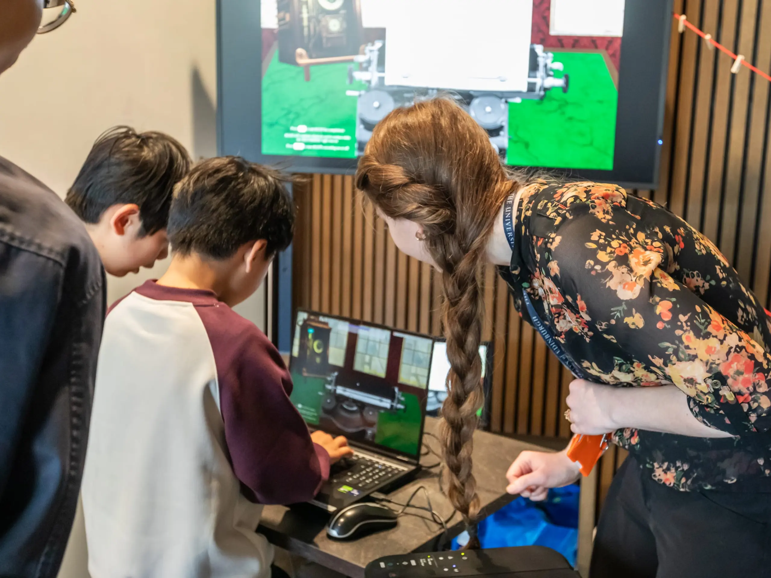 A woman with a long plait helps two boys using a laptop, with a large monitor displaying a computer game or simulation in the background. They are in an indoor setting with wooden panelling.