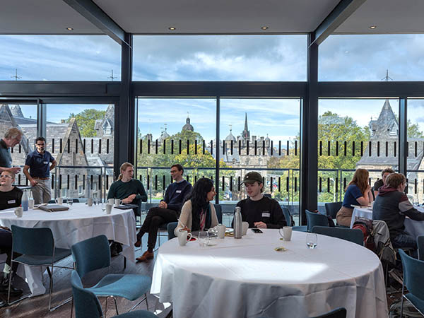 People sit and converse at round tables in a bright room with large windows overlooking historic buildings and greenery. Some stand and talk, whilst others sit, with coffee pots and glasses on the tables.