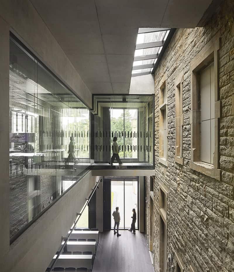 A modern building interior features glass walkways above a stone wall, with natural light streaming in through skylights. Two people stand near the entrance, and one person is seated on the upper walkway.