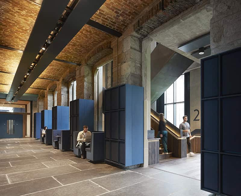 A modern office space with exposed brick ceilings, stone walls, blue cubicles, and large windows. People are working at desks and walking near a wooden staircase marked with the number 2.