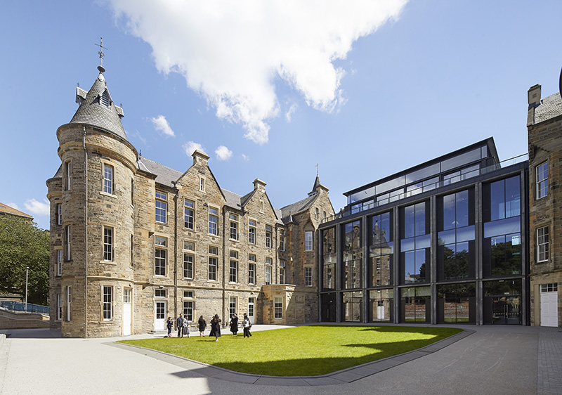 A historic stone building with turrets stands next to a modern glass extension. Several people are gathered on a grassy courtyard under a blue sky with scattered clouds.