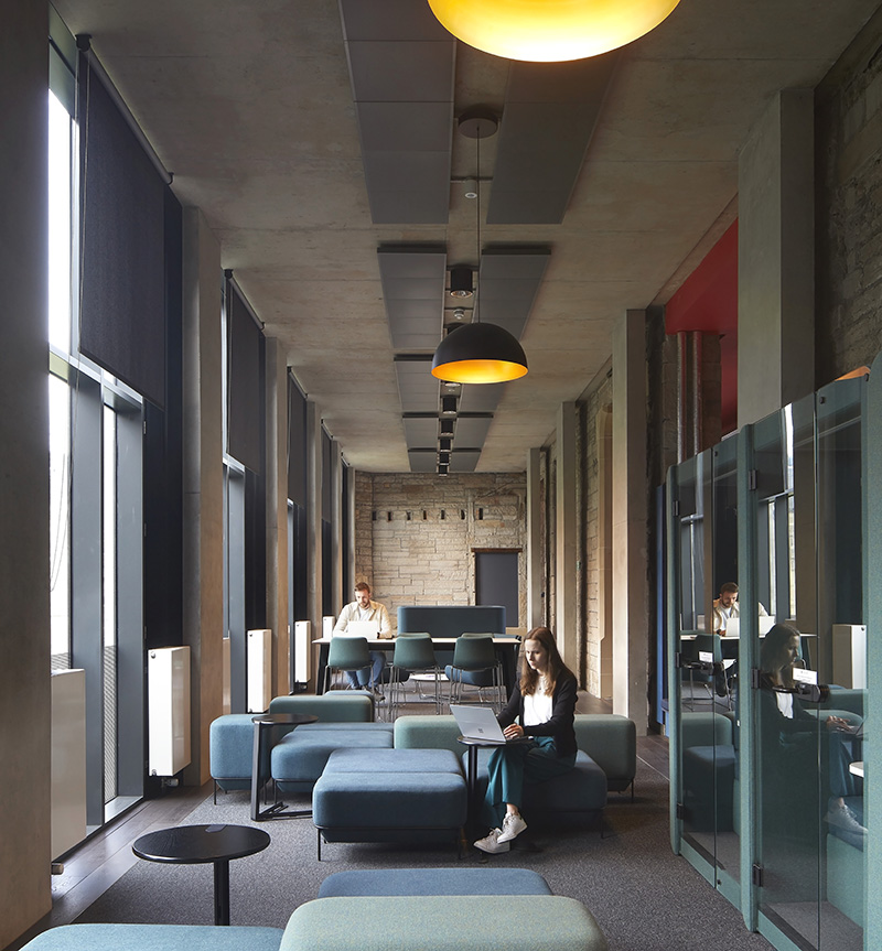 Modern lounge area with large windows, green and blue seating, overhead pendant lights, and people working on laptops at tables and sofas. The space feels open, bright, and contemporary.