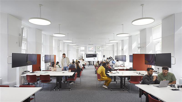 Bright, modern office space with large windows, round ceiling lights, and white walls. People work in groups at desks with computers. Some collaborate, whilst others work individually. The atmosphere appears busy and focused.
