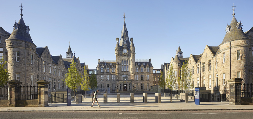 A person walks past Edinburgh Futures Institute, a grand, historic stone building with turrets, spires, and a central clock tower under a clear blue sky. The entrance is framed by young trees and a gated courtyard.