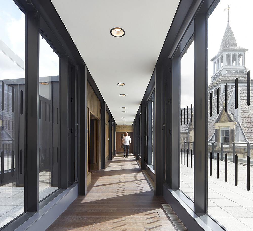 A modern glass corridor with wooden floors and black frames, featuring ceiling lights. A person stands in the distance. Outside, through the windows, a historic stone building with a clock tower is visible.