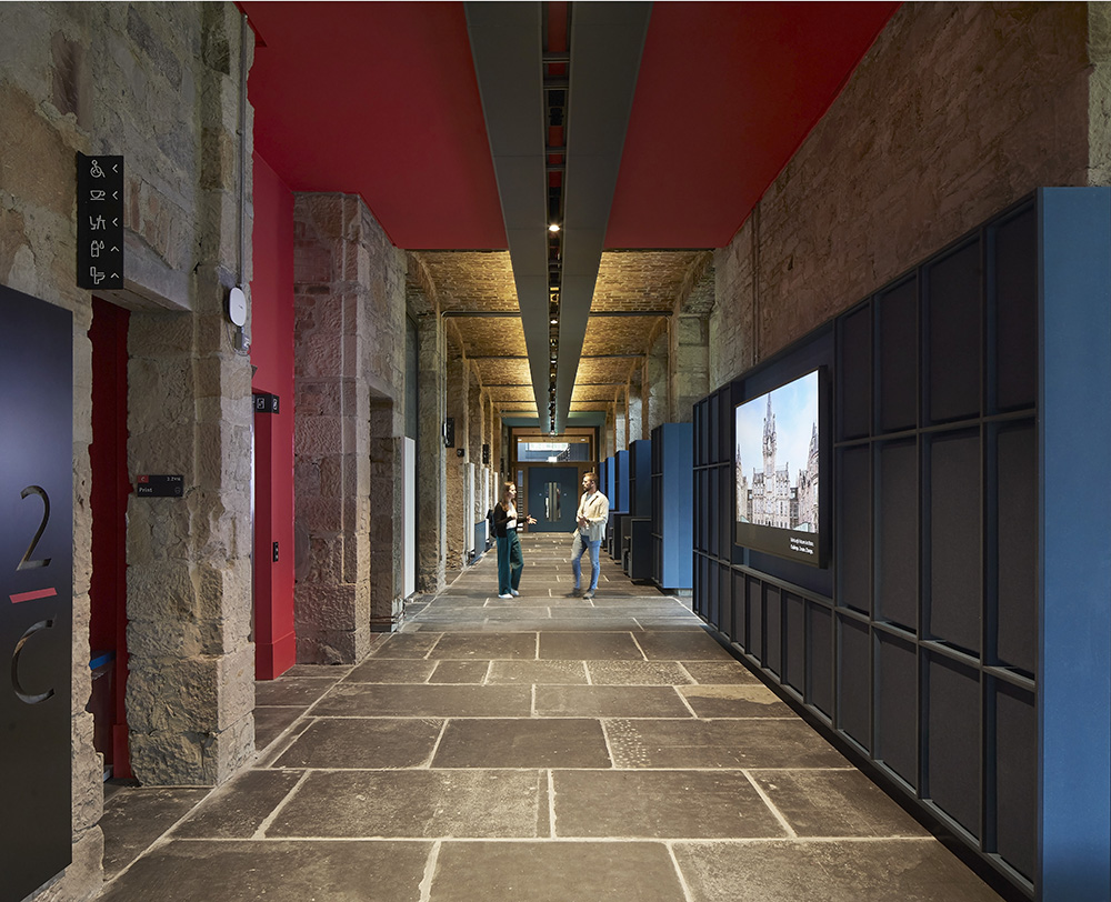 A modern corridor with stone walls and flooring, red ceiling accents, black doors, and digital screens. Two people are walking down the corridor, which is well-lit by ceiling lights.
