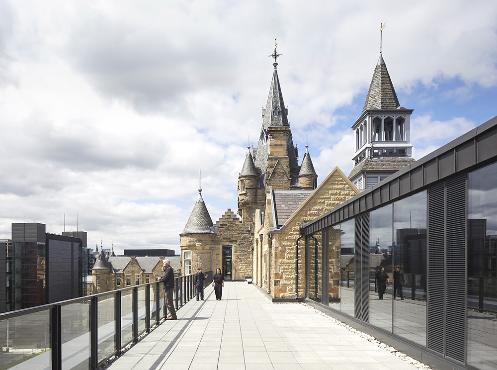 A rooftop terrace with glass balustrades overlooks a historic stone building with towers and spires; people stand along the edge, and modern glass buildings are visible in the background under a partly cloudy sky.