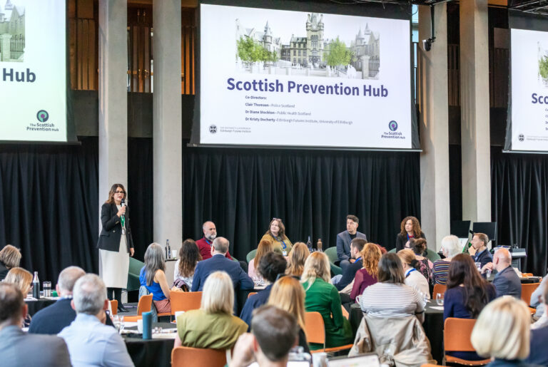 A woman stands speaking at a lectern while four panellists sit onstage in front of an audience. Two large screens display "Scottish Prevention Hub" with images of buildings and event branding.
