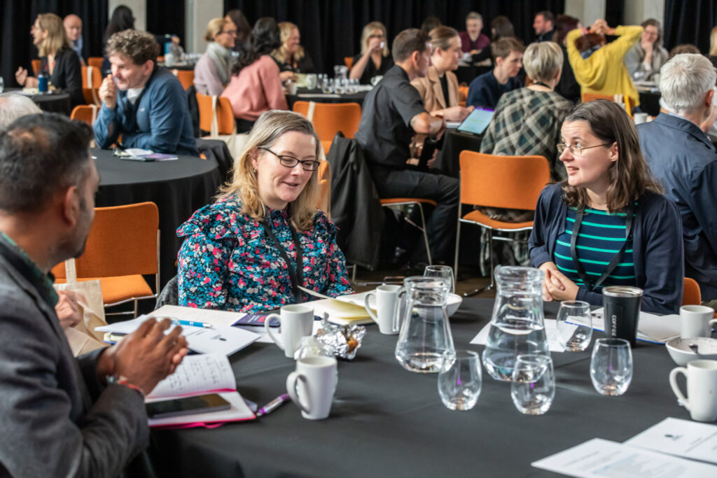 A group of people sit around round tables, engaged in conversation at a conference or workshop. Papers, glasses, mugs, and water jugs are on the tables. The room is busy with attendees talking in small groups.
