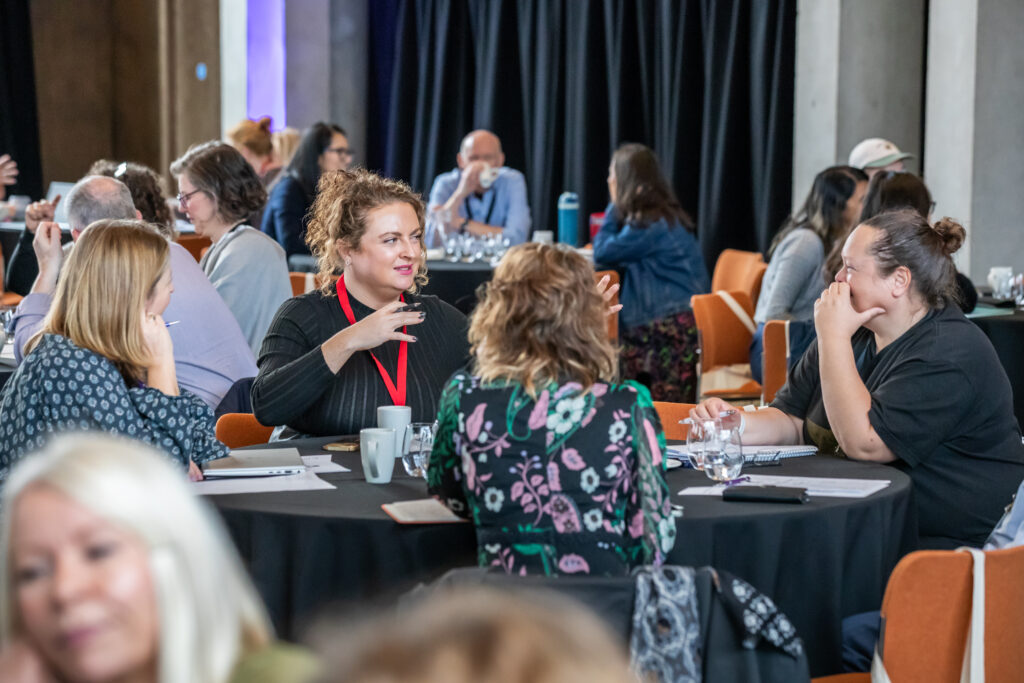 People sit in groups around round tables, talking and engaging in discussion at a conference or workshop. The atmosphere appears lively and collaborative, with notebooks, cups, and water glasses on the tables.