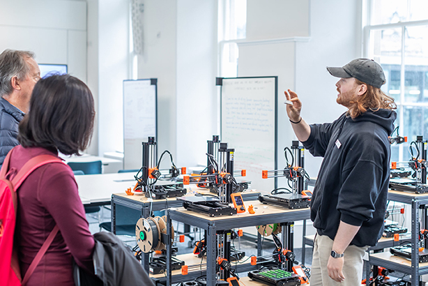 A man in a black cap explains something to two people in a room filled with 3D printers and whiteboards, suggesting a workshop or classroom setting with technical equipment.