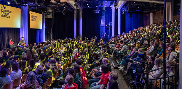 A large audience sits in a modern auditorium, watching a speaker on stage at the Edinburgh Futures Institute event. Two large yellow screens display the event’s name. The room is brightly lit and filled with attentive attendees.