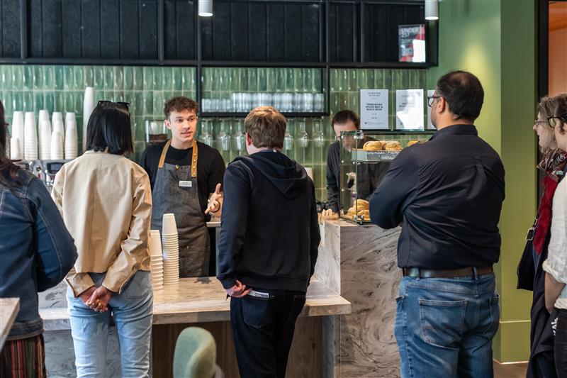 A barista behind a counter talks to a group of customers in a modern café. Glasses, cups, and pastries are visible on shelves and in a display case. The customers queue, waiting to order.