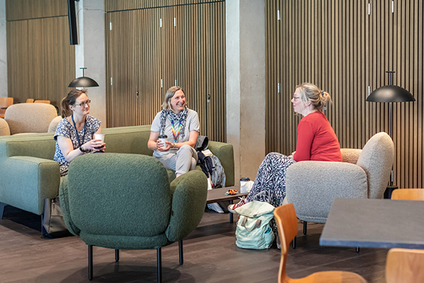 Three people sit on cushioned chairs in a modern lounge, talking and smiling. A rucksack and some cups are on the table between them. Wooden panelling and soft lighting are in the background.