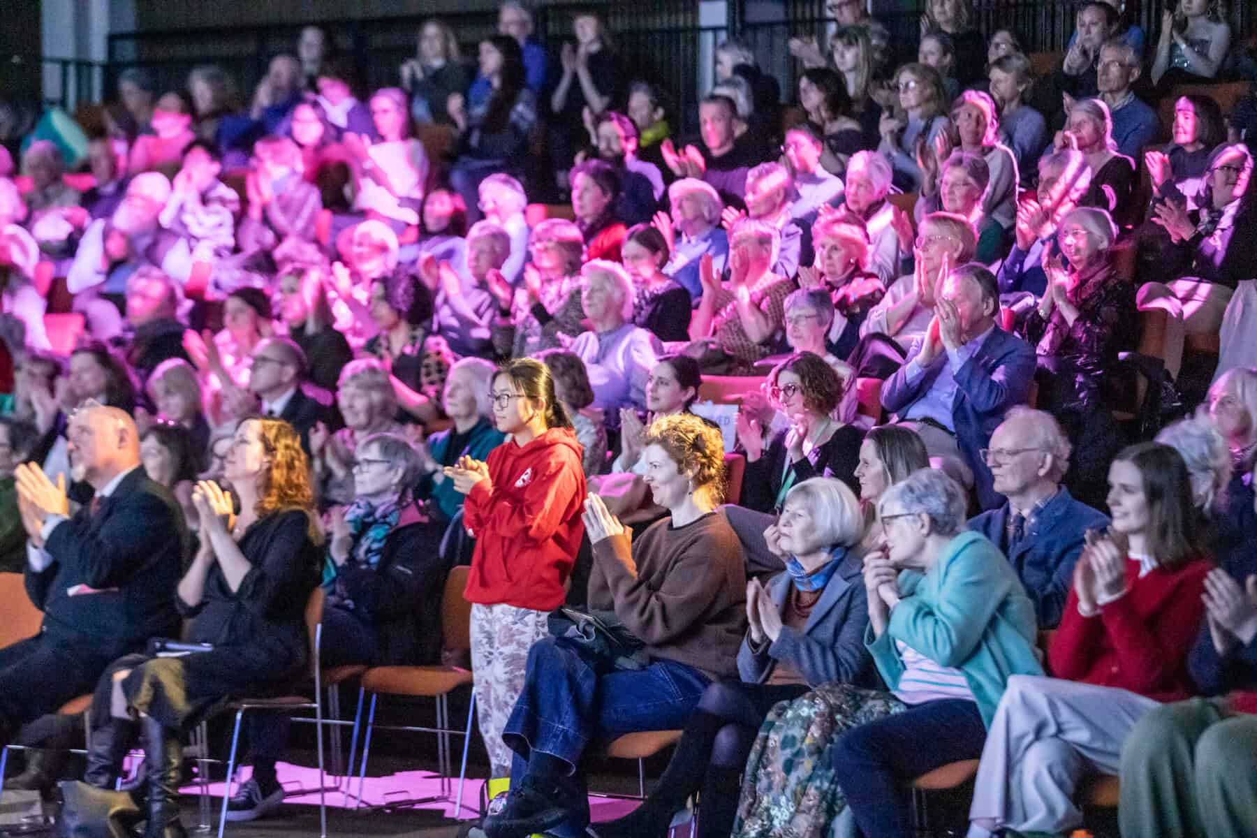 A large audience sits in tiered seating, clapping and watching an event. The lighting is colourful with shades of purple and pink, creating a lively atmosphere. People of various ages are visible, some standing and applauding.