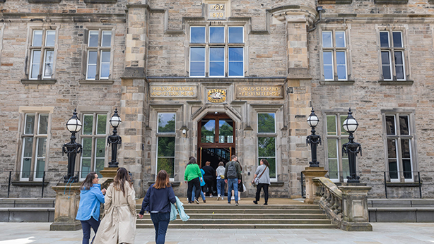 Several people walk towards the entrance of a historic stone building with large windows, ornate lamps, and a sign above the doorway.