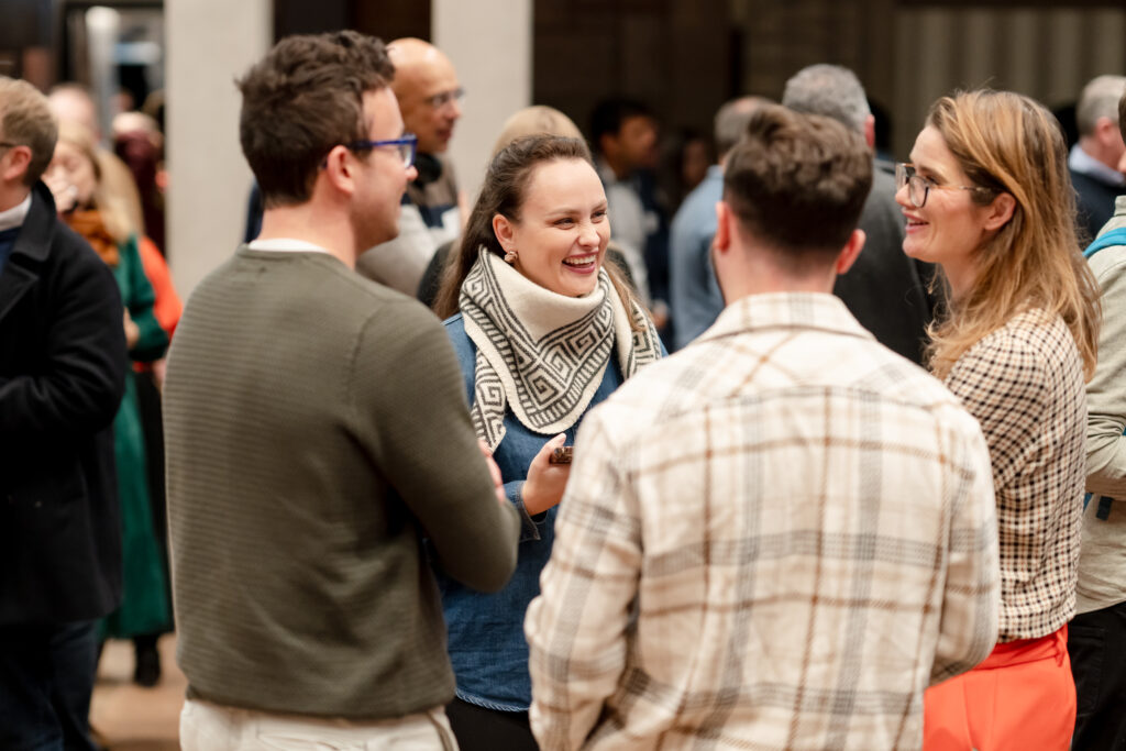 Four people stand in a group, smiling and talking at an indoor social event. Other attendees can be seen mingling in the blurred background. The setting appears warm and lively.