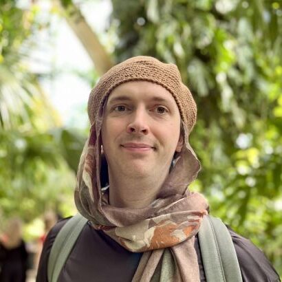 A person wearing a brown knitted hat and patterned scarf stands outdoors in front of lush, green foliage, smiling gently at the camera.