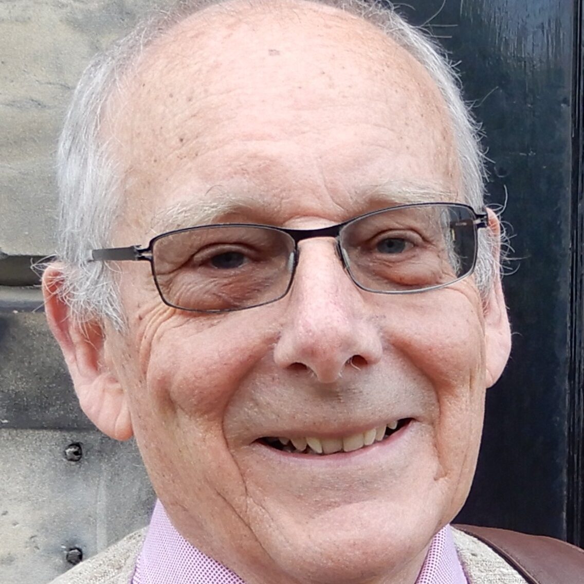 An older man with short grey hair and glasses smiles at the camera. He is wearing a light-coloured jacket, a pink shirt, and a blue tie, standing in front of a stone wall and a black door.