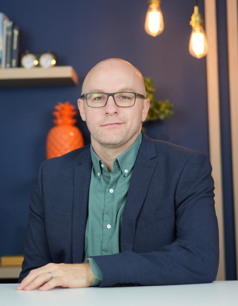 A man with glasses and a shaven head, wearing a green shirt and dark blazer, sits at a white table. Behind him are a shelf with books and an orange pineapple, a plant, and two hanging lights against a blue wall.