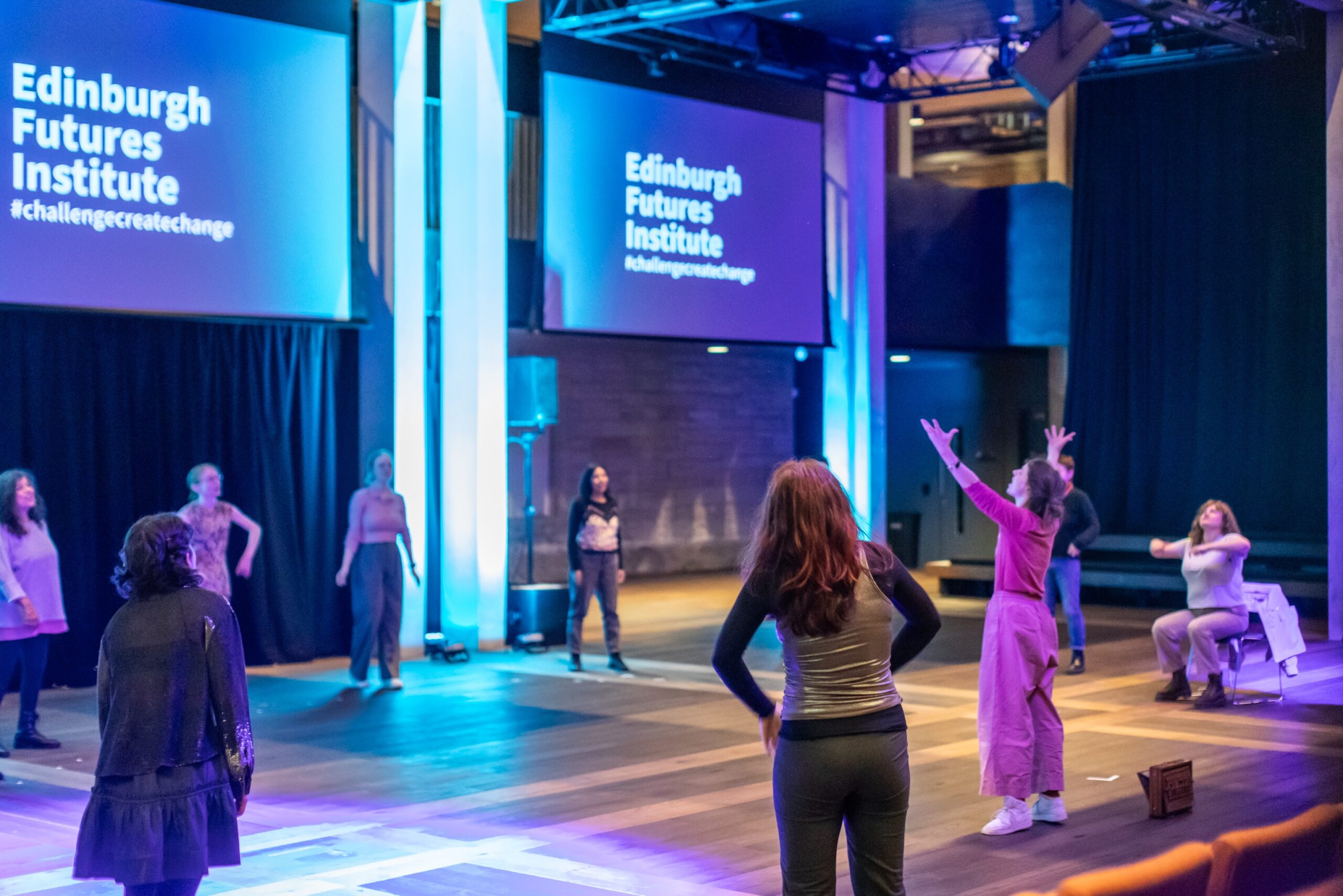 A group of people stand in a spacious, modern room with wooden floors, participating in an interactive workshop. Large screens display "Edinburgh Futures Institute" and "#challengecreatechange".