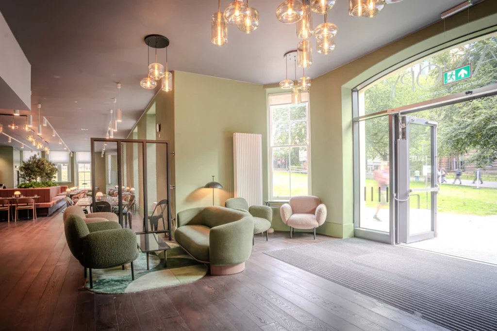 Modern lounge area with green walls, round glass pendant lights, plush green and beige chairs, large windows, and open glass doors leading outside. Natural light brightens the room, and people can be seen outside.