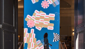 A person walks past a large, colourful digital screen displaying abstract flower and geometric shapes on a blue background inside a building with tall open doors.