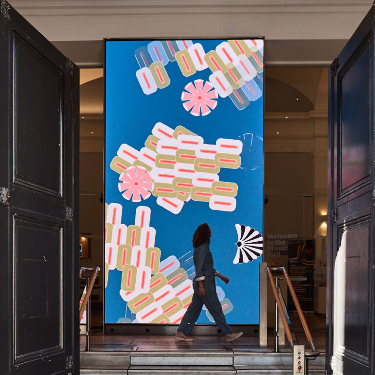 A person walks past a large, colourful digital screen displaying abstract flower and geometric shapes on a blue background inside a building with tall open doors.