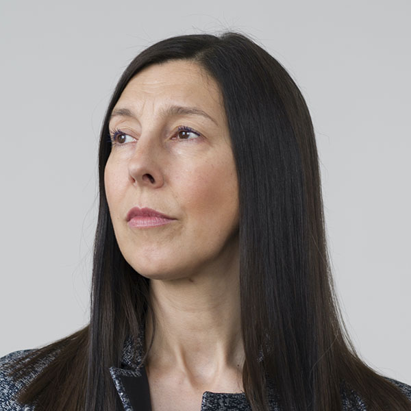 A woman with long straight dark hair and light skin is wearing a textured dark jacket. She is looking slightly to the left with a neutral expression against a plain light grey background.