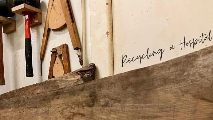 A close-up of a workshop wall with woodworking tools, a plank of reclaimed timber, and the words "Recycling a Hospital" handwritten on a white surface.