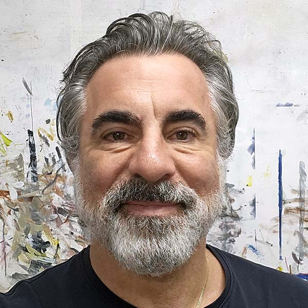 A middle-aged man with grey hair and a beard smiles at the camera, standing in front of a light-coloured, abstract painted background.