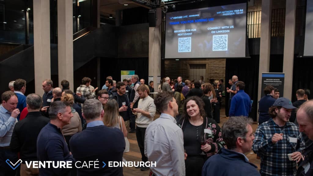 A large group of people socialises and networks at an indoor event. Two screens display messages about registering for the next event and connecting on LinkedIn. "Venture Café Edinburgh" is displayed in the corner.
