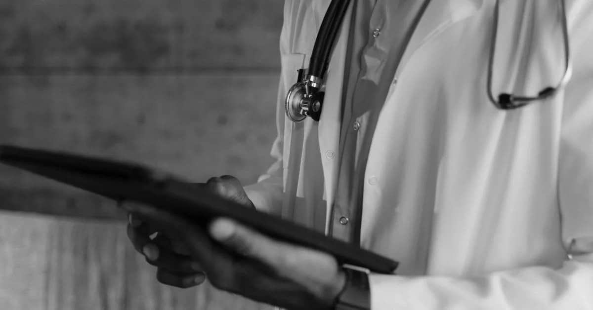 A doctor wearing a white coat and stethoscope holds and uses a digital tablet. The image is in black and white, and the doctor's face is not visible.