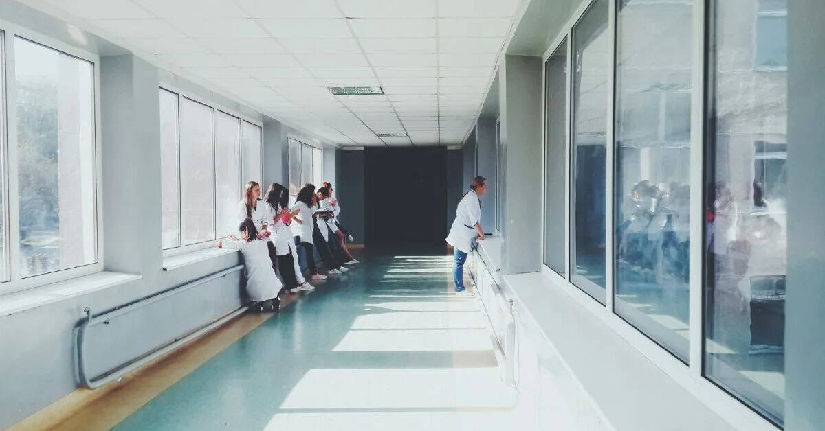 A group of people in white lab coats stand and talk near windows in a bright hospital corridor, whilst one person walks alone towards the end of the corridor.