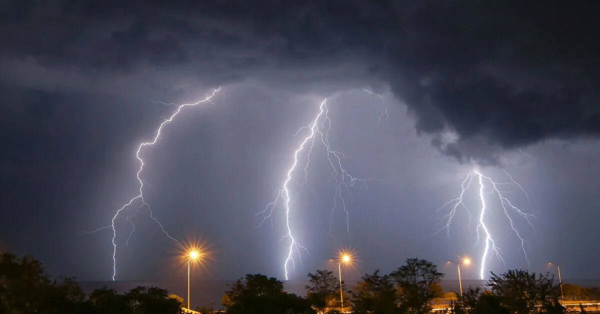 Three bright lightning bolts strike down from dark storm clouds above a nighttime landscape. Streetlights illuminate trees and buildings below, contrasting with the dramatic, illuminated sky.