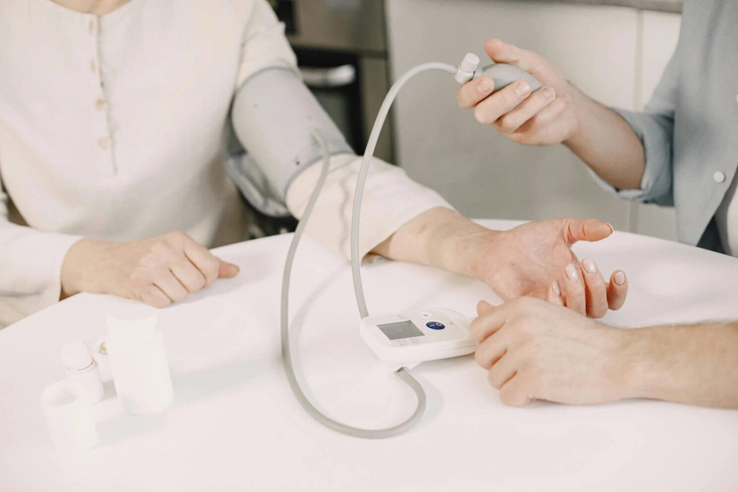 A healthcare professional measures a patient's blood pressure using an electronic monitor at a table. The patient's arm is in a cuff, and medicine bottles are visible on the table.