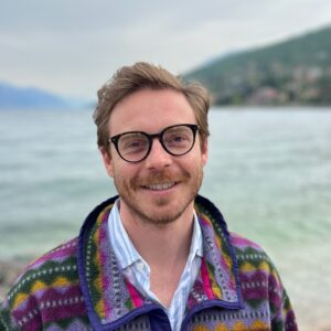 A smiling man with glasses and a beard stands outdoors by a body of water, wearing a colourful patterned jacket and a striped shirt. Blurred green hills and a cloudy sky are in the background.