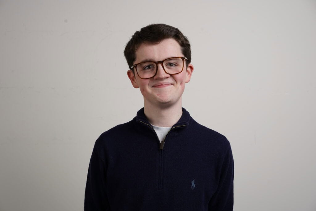 A young person with short brown hair and glasses smiles at the camera. They are wearing a navy blue quarter-zip jumper and standing in front of a plain white background.