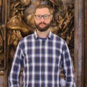 A man with short hair, glasses, and a beard wears a blue and white checked shirt, standing in front of an ornate, bronze-coloured sculpted background.