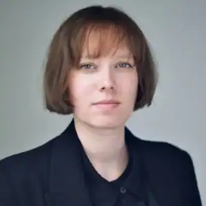 A person with short, straight light brown hair and a fringe, wearing a black blazer over a black shirt, looks directly at the camera with a neutral expression against a plain light background.