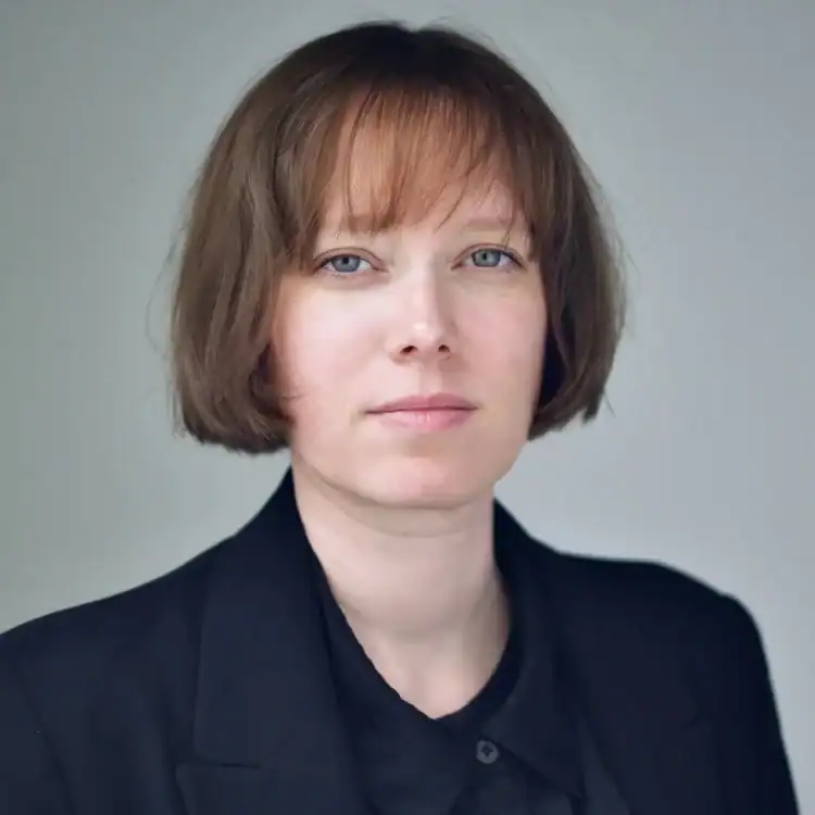 A person with short, straight light brown hair and a fringe, wearing a black blazer over a black shirt, looks directly at the camera with a neutral expression against a plain light background.