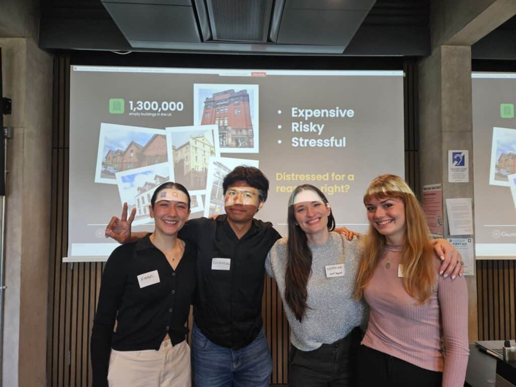 Four young adults wearing name badges and headbands stand smiling in front of a presentation slide about the property market. The slide lists "Expensive, Risky, Stressful" and shows images of buildings.