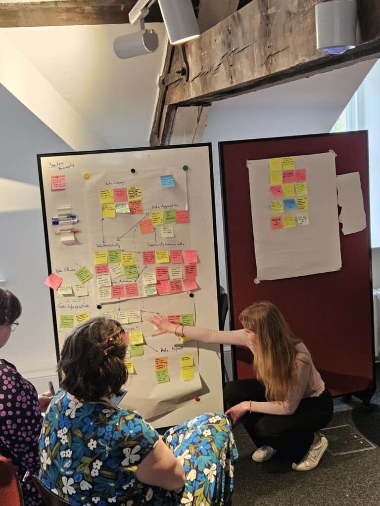 A woman kneels and gestures towards a large whiteboard covered with colourful sticky notes, while two women seated in front listen attentively. Another board with more sticky notes is in the background.