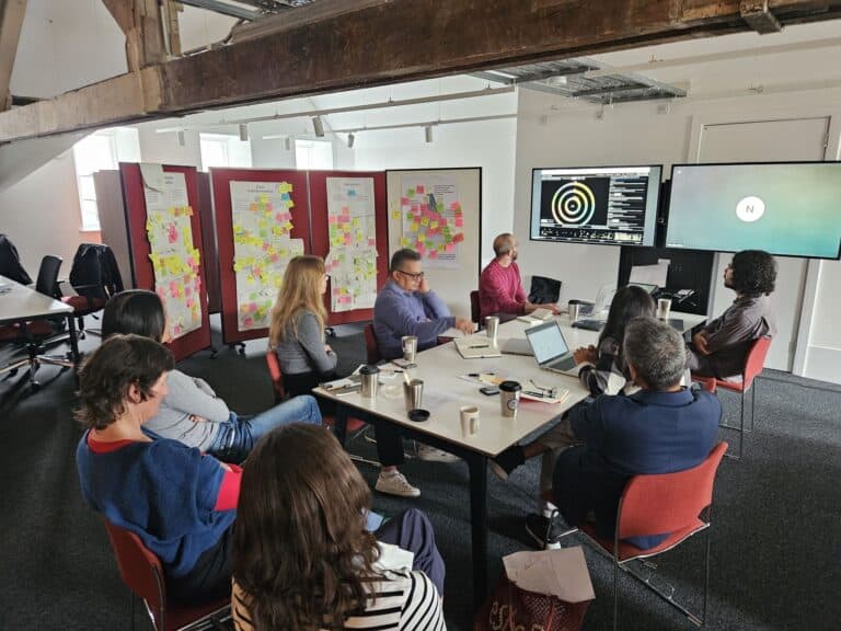 A group of people sit around a table in a meeting room, with laptops, papers, and coffee cups. Large boards covered in colourful sticky notes and a presentation screen are visible in the background.