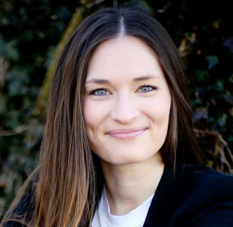 A woman with long straight brown hair and blue eyes smiles at the camera. She is wearing a black blazer and a white top, with a blurred outdoor background of greenery.